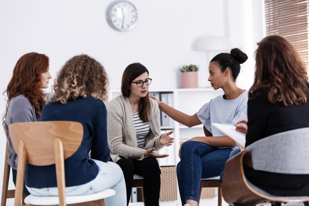 Group therapy in a clean, modern office is shown as five women sit in a semi-circle of wooden chairs, actively listening and sharing.