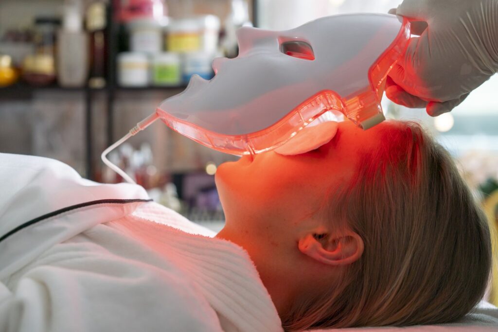 Red light therapy is shown with a gloved technician holding a bright red LED face mask over a client's face in profile, casting a warm red glow.