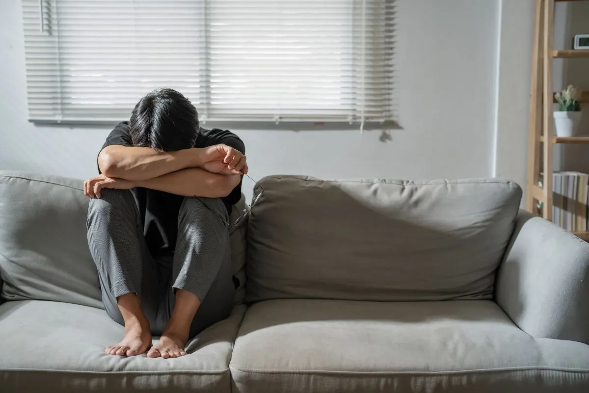 An anxiety disorder is depicted as a silhouetted figure sitting on a couch with their head buried in their hands in a simple room.