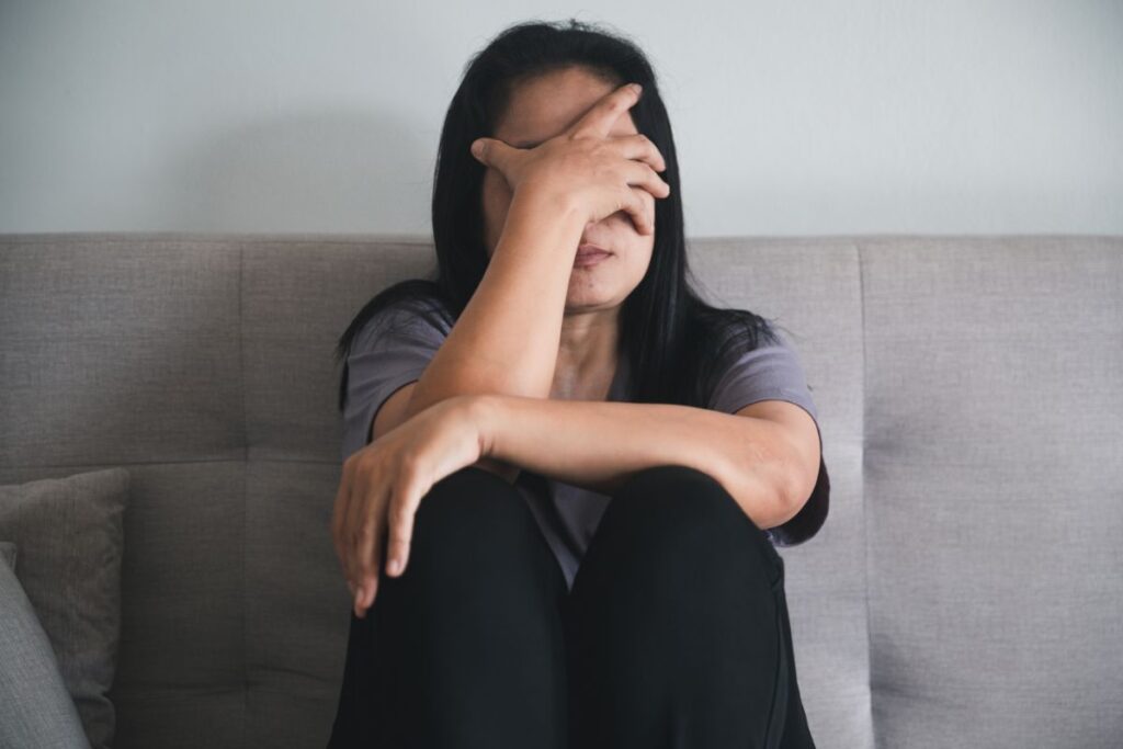 A young woman who is struggling with mental health sitting on a couch holding her face in her hands.