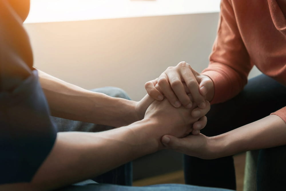 Mental health support is shown through a close-up of two people holding hands during a therapy session to provide comfort and connection.