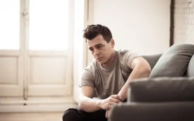 Post traumatic stress disorder is shown by a man sitting on the floor leaning against a grey couch with a somber and downcast expression.