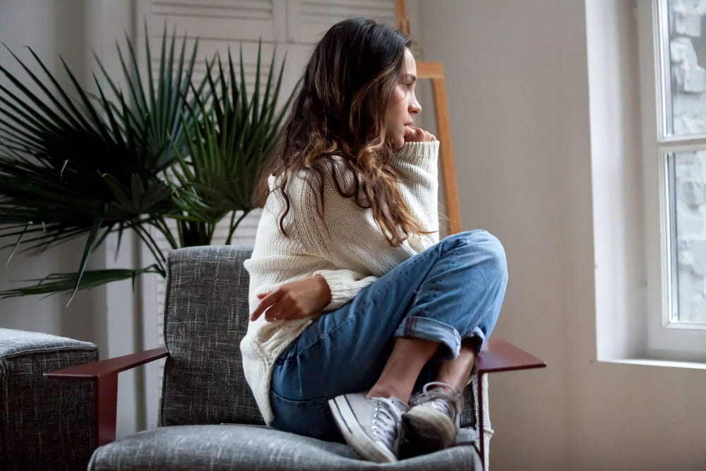 A woman sits contemplatively in a chair at a schizophrenia treatment center while looking toward a window in a bright, sunlit room.