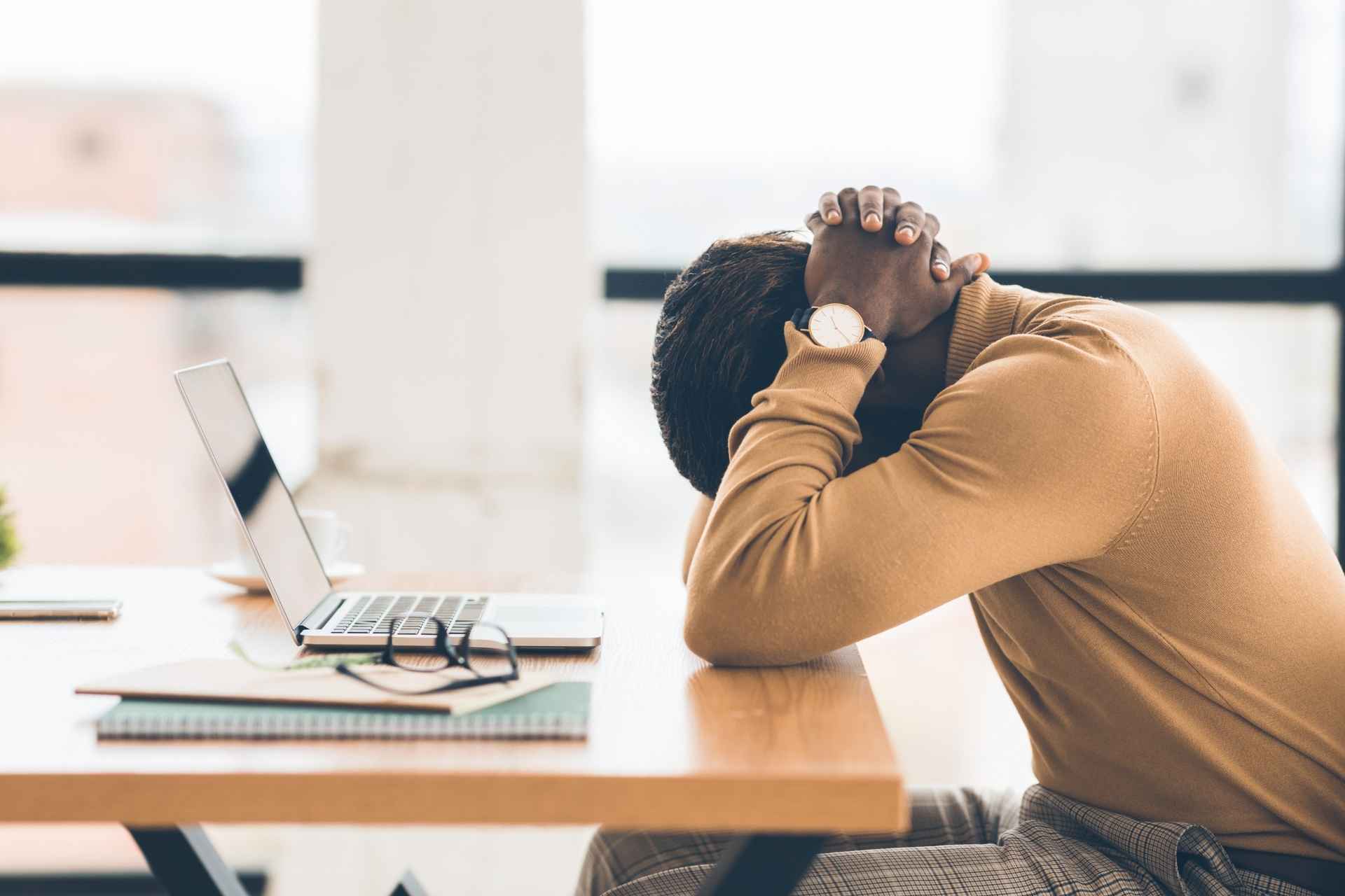 Exhausted employee sitting at a desk with his head down beside a laptop.